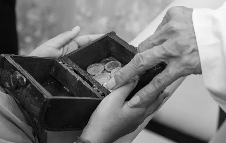 Black and white close-up of two pairs of hands over a small open wooden box of coins, one hand passing a coin to the other