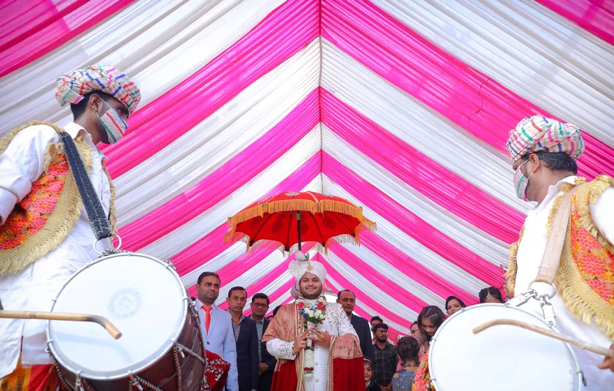 Two drummers in white with large barrel drums flank a groom in a turban walking under a red umbrella beneath a pink and white striped canopy