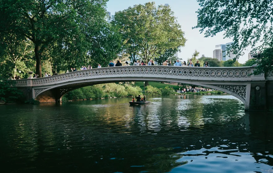 Bow Bridge in Central Park lined with people along its railing, a rowboat on the Lake below and trees on both banks