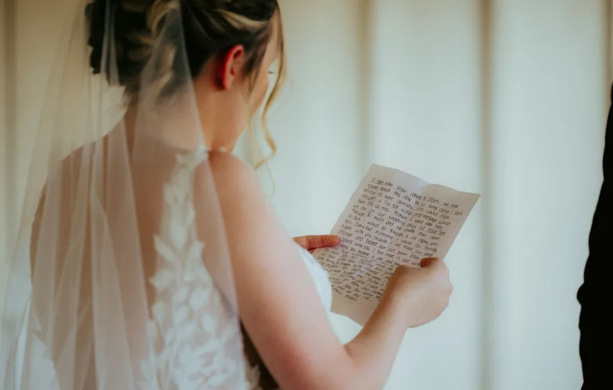 A bride in a lace dress and veil, seen from behind, reads a handwritten letter on lined paper