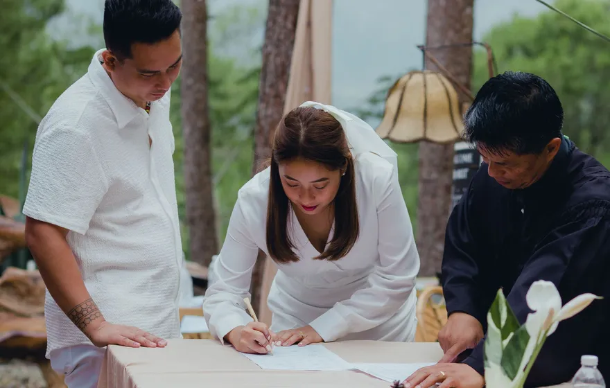 A bride in white leans over a table to sign a marriage certificate while the groom and the officiant stand beside her outdoors