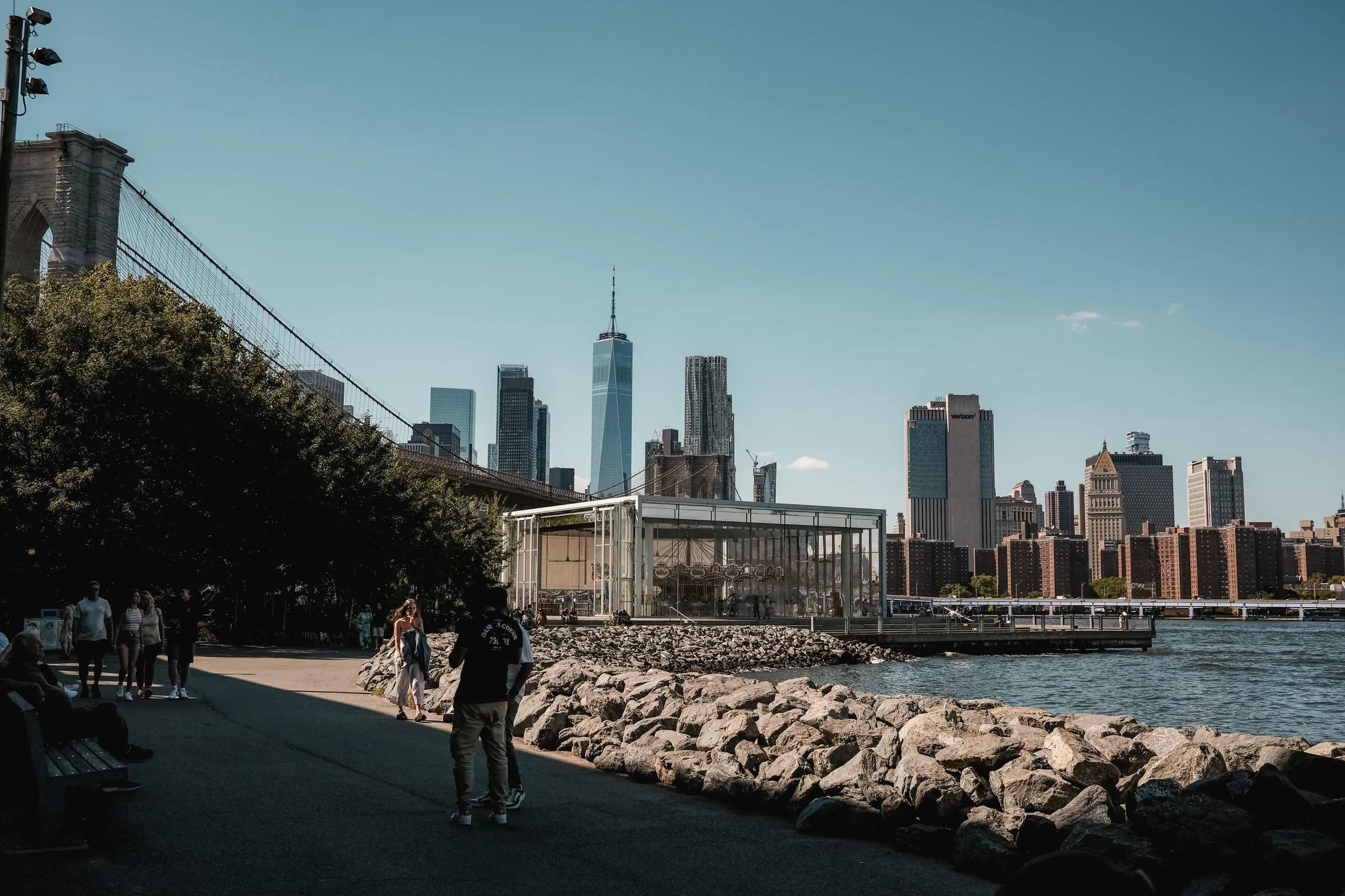 The waterfront path in Brooklyn Bridge Park beside the Brooklyn Bridge, with the glass pavilion of Jane's Carousel and the Lower Manhattan skyline across the East River
