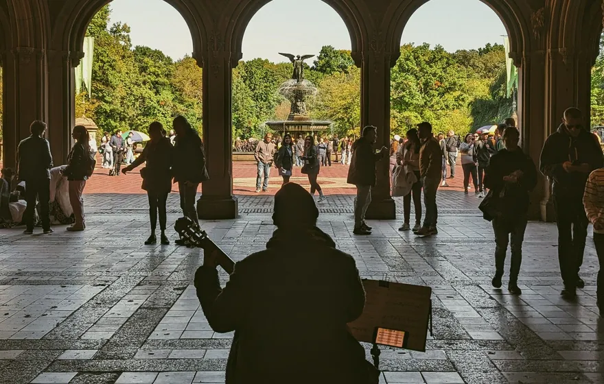 A guitarist sits in silhouette under the tiled ceiling of the Bethesda Arcade, music stand in front of him, the fountain visible through the arches beyond