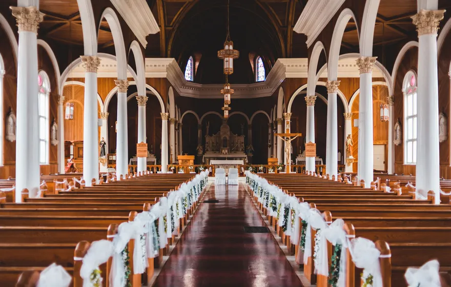 The center aisle of a large church with white columns and arches, wooden pews tied with white ribbon and flowers leading to the altar