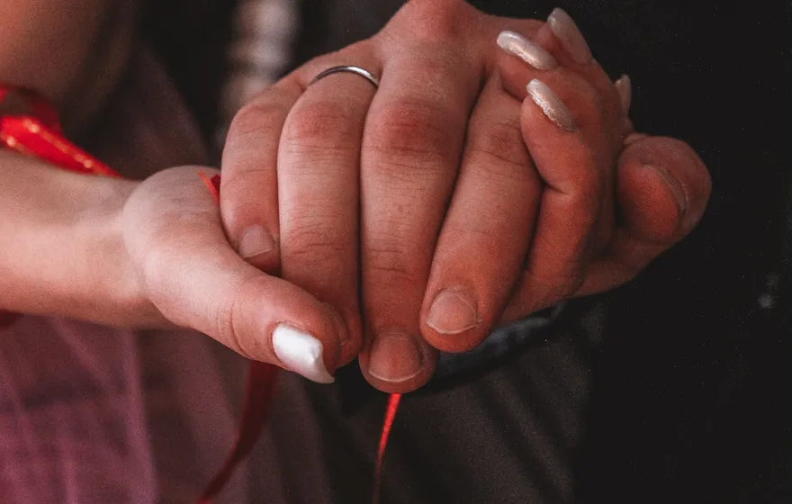 Close-up of a couple's clasped hands, a wedding band showing, with a thin red ribbon trailing down from their joined hands