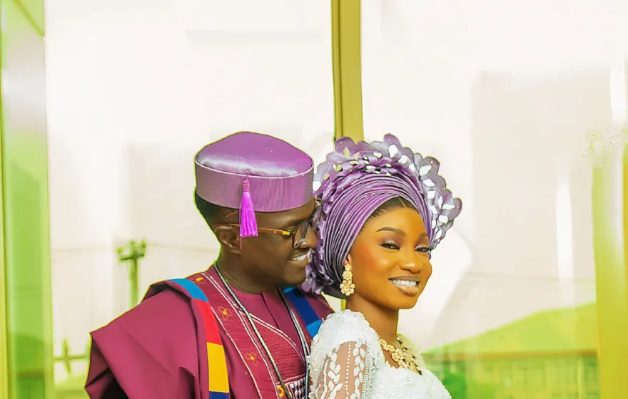 A smiling couple in Nigerian traditional wear, the groom in a magenta agbada and cap, the bride in a lilac gele head wrap and white lace