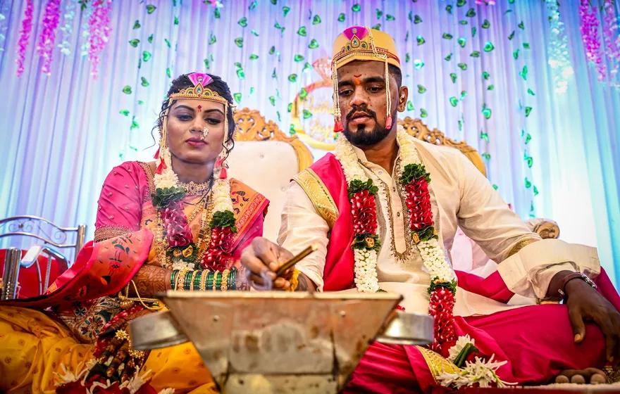 A bride in a pink sari and gold headpiece and a groom in a cream kurta and embroidered cap, both in red and white flower garlands, sit before a metal fire vessel as the groom holds a small stick over it