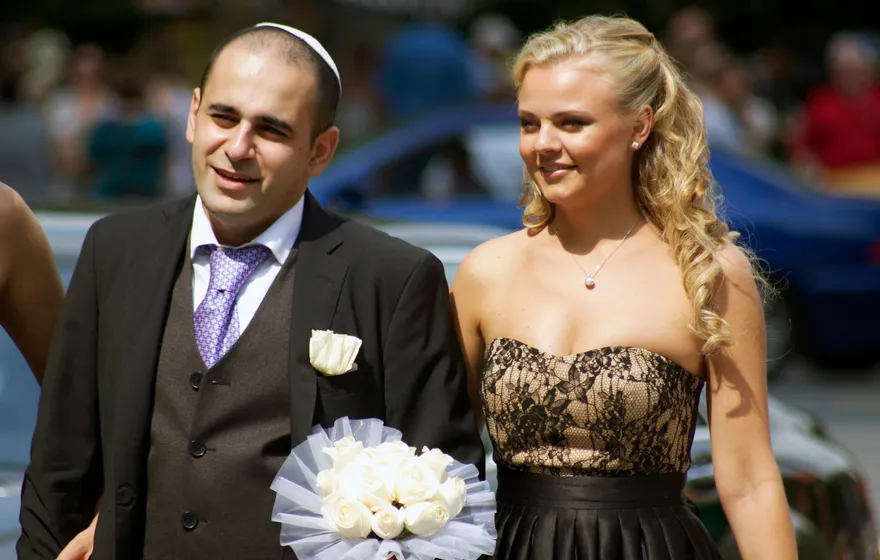 A smiling groom in a white kippah and a gray suit walks beside his bride, who holds a bouquet of white roses, on a Central Park path