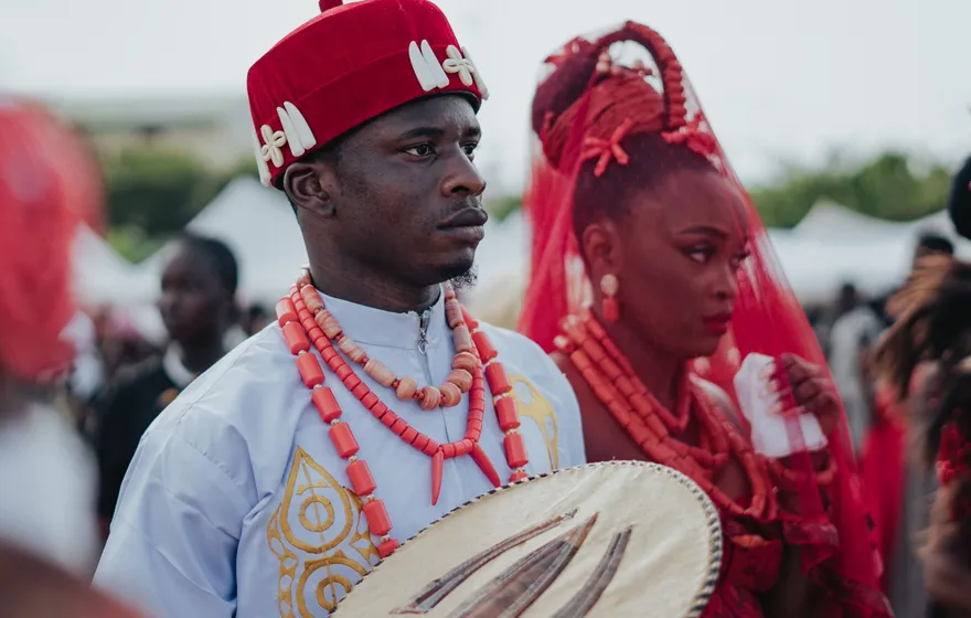 A groom in a red cap and coral bead necklaces stands beside a bride in a red veil and coral beads, guests and tents behind them at an outdoor Nigerian wedding