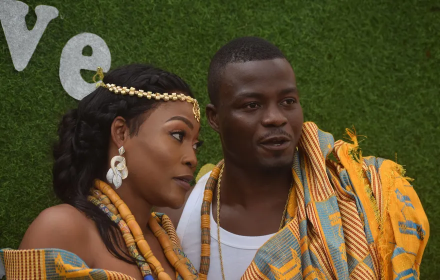 A couple in kente cloth and coral bead necklaces sit close together, the bride in a gold headpiece turning toward the groom