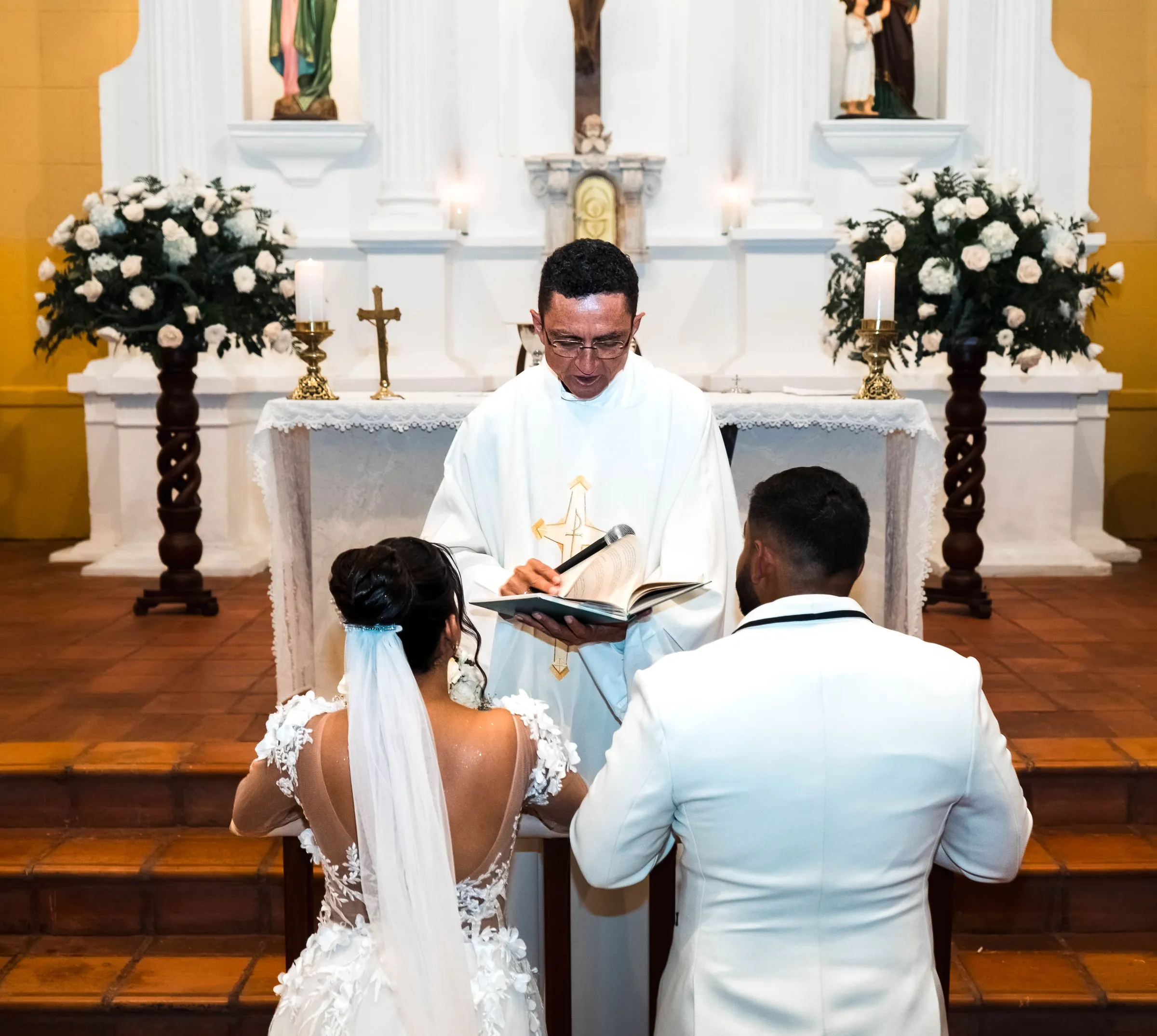 A bride and groom kneel at a church altar while a priest in white vestments reads from a book, a crucifix and white flowers behind him