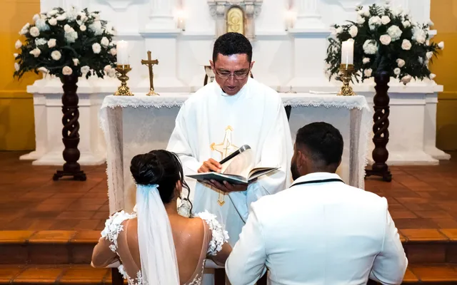 A bride and groom kneel at a church altar while a priest in white vestments reads from a book, a crucifix and white flowers behind him