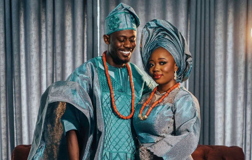 A smiling couple in matching teal and silver traditional Nigerian outfits, the bride in a gele and both wearing coral beads