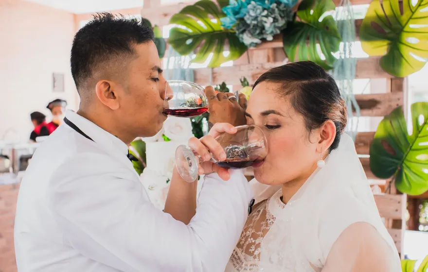 A groom in a white jacket and a bride in a veil link arms and drink red wine from their glasses at the same moment