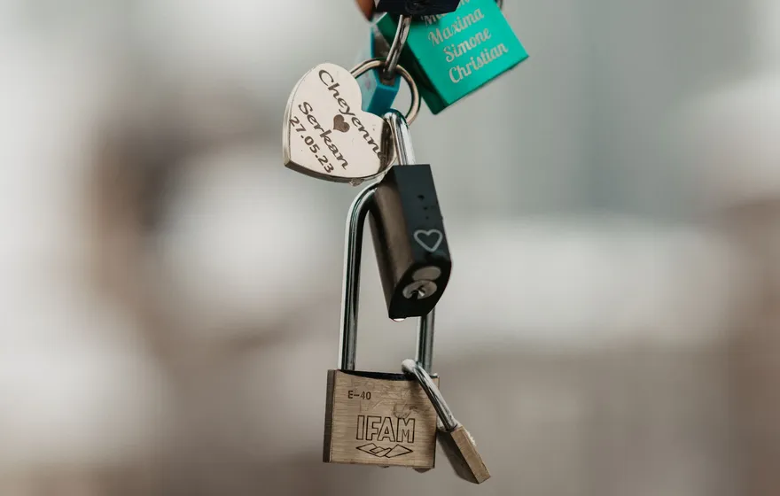 Several engraved padlocks hooked together on one ring, including a silver heart-shaped lock engraved with two names and a date and a green lock engraved with names
