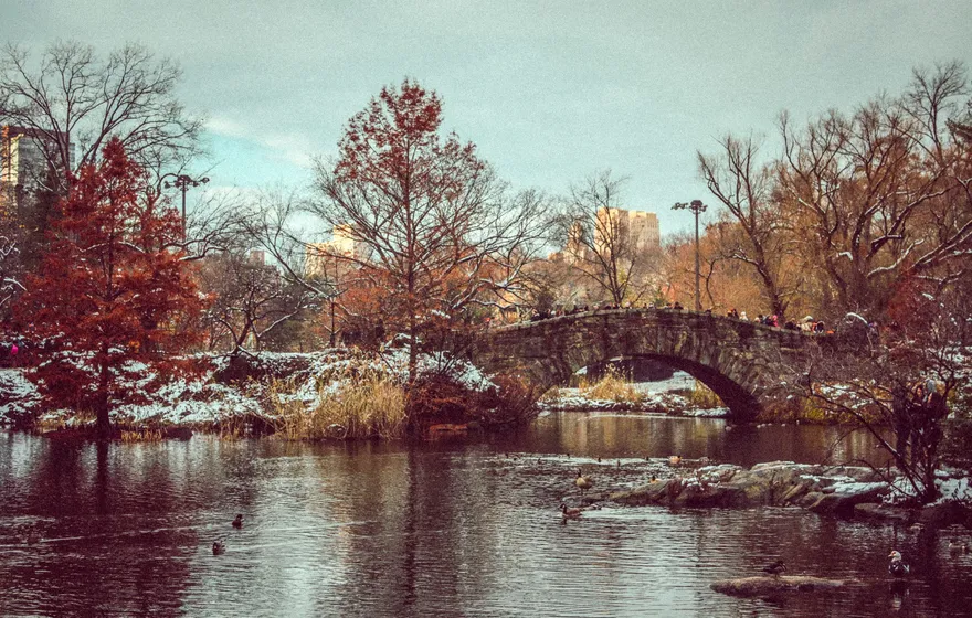 Gapstow Bridge with a light covering of snow on its stone and the banks, bare red and brown trees around the Pond and apartment towers behind