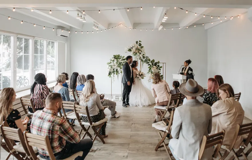A small group of guests on wooden chairs watches a couple stand under a floral arch while an officiant reads at a lectern in a bright white room
