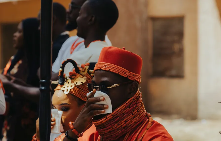 A man in a red cap and coral beads sips from a small cup at an outdoor Nigerian wedding, a woman in a beaded headpiece beside him and guests behind