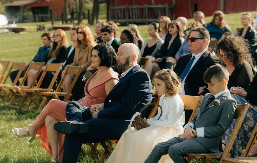 Wedding guests of all ages, including two children, sit in rows of wooden folding chairs on a lawn, watching a ceremony