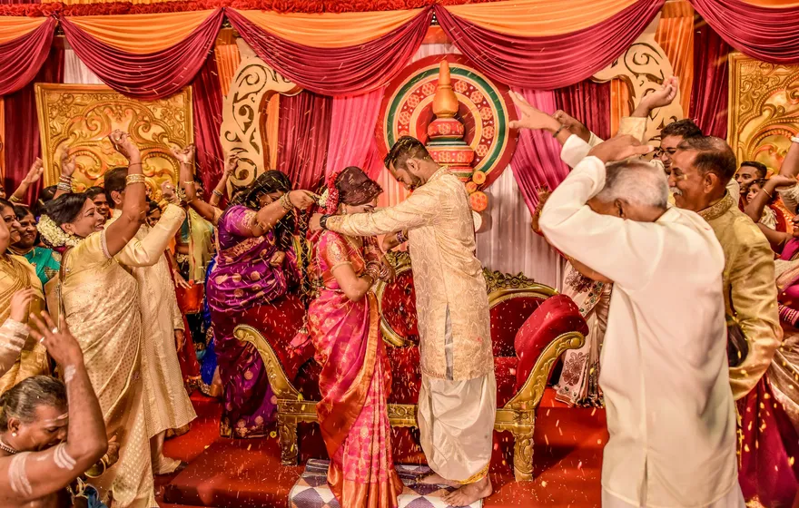 A groom in a cream sherwani reaches to the neck of a bride in a pink silk sari on a stage hung with red and gold drapes, while family members on both sides throw yellow rice over them