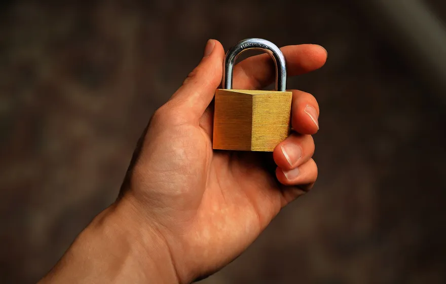 An open hand holding a small brass padlock with a steel shackle against a dark brown background