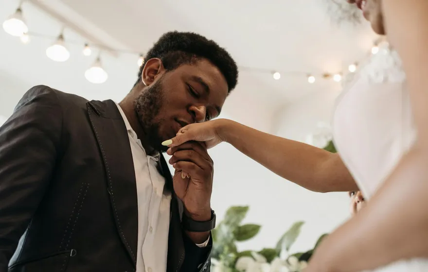 A groom in a dark jacket gently kisses the back of his bride's hand during their wedding, warm string lights glowing softly behind them