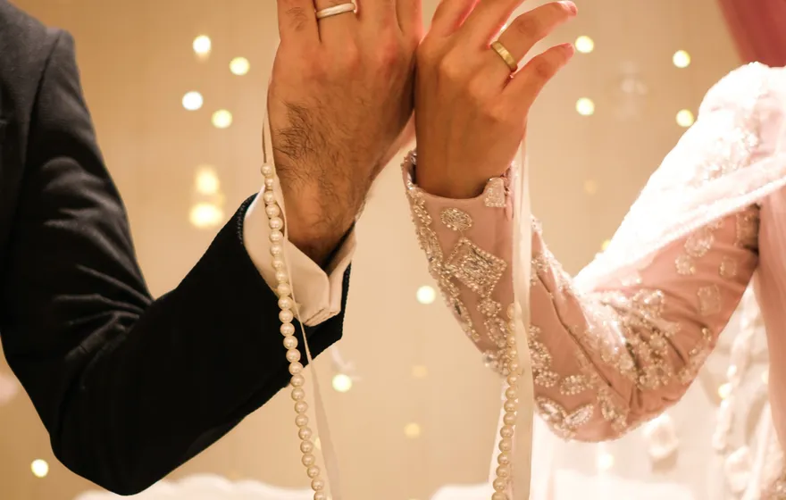 A groom's and a bride's raised hands, both wearing wedding bands, joined at the wrist by a strand of pearls and a white ribbon hanging below