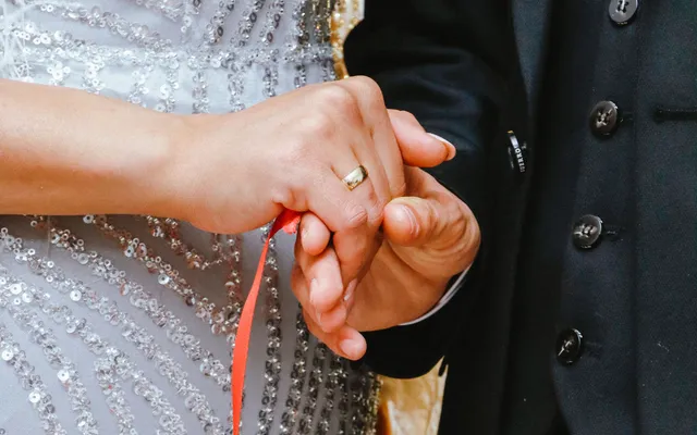 Close-up of a bride and groom's joined hands with a red ribbon wrapped around them, her gold wedding band showing