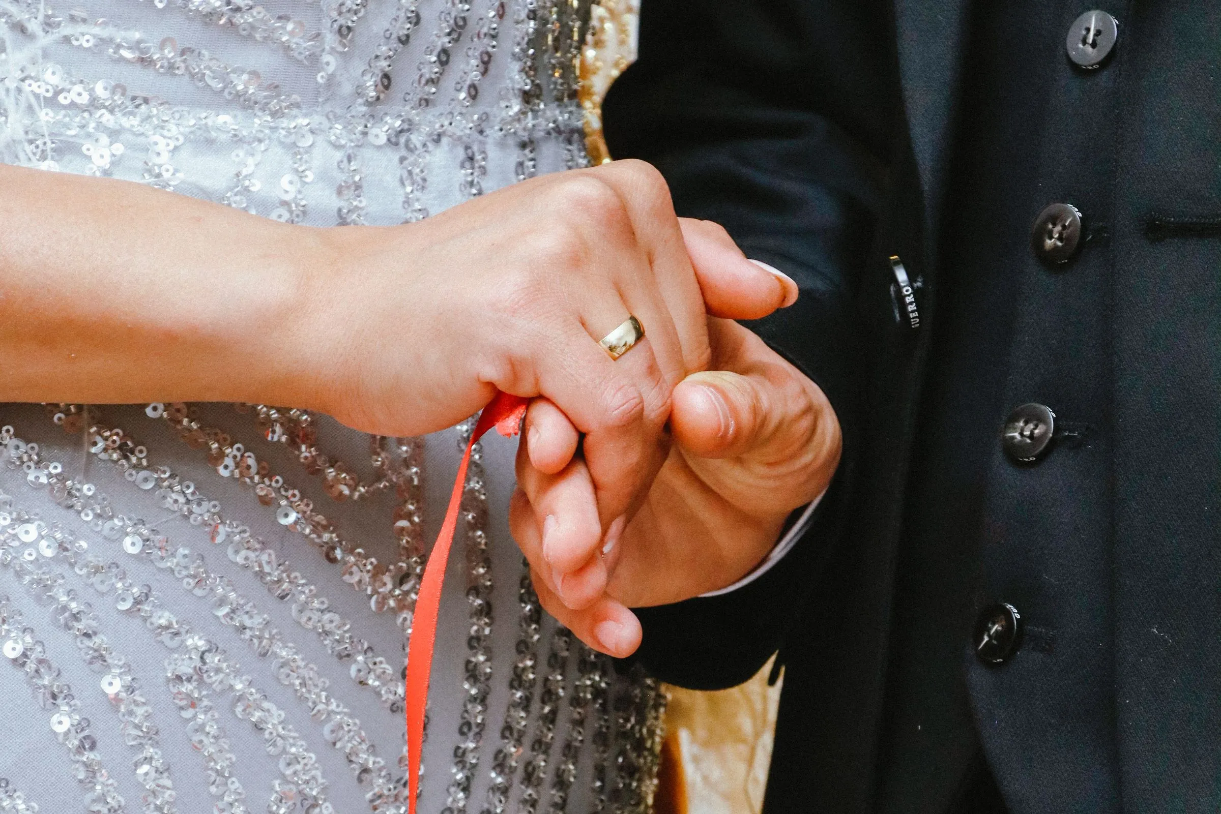 Close-up of a bride and groom's joined hands with a red ribbon wrapped around them, her gold wedding band showing