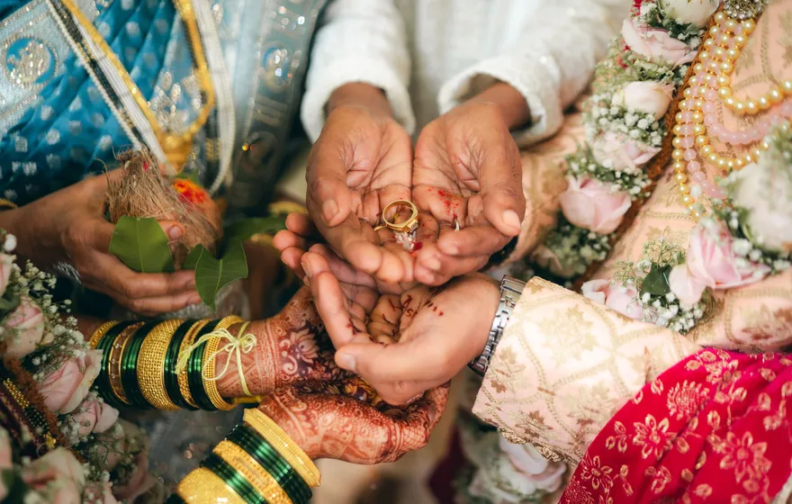 Close-up of hennaed hands in gold bangles and bright silk cupped together around two rings during a traditional Indian wedding ritual