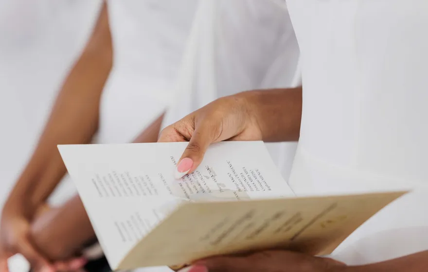 Close-up of a woman's hands holding open a folded cream ceremony program printed with lines of text, a white dress behind it