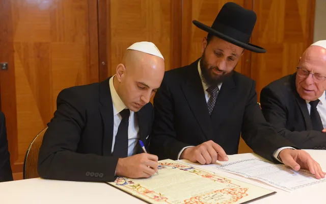 A man in a white kippah signs an illuminated Hebrew document at a table while a man in a black hat and an older man in a kippah look on