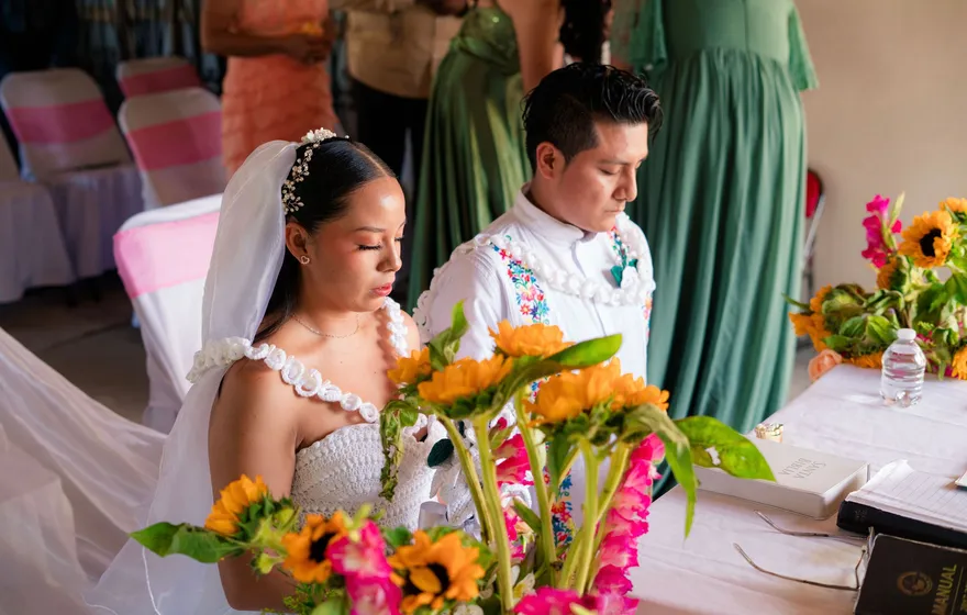 A bride in a veil and a groom in an embroidered white shirt sit with heads bowed at a ceremony table of sunflowers, a white corded lazo resting across their shoulders