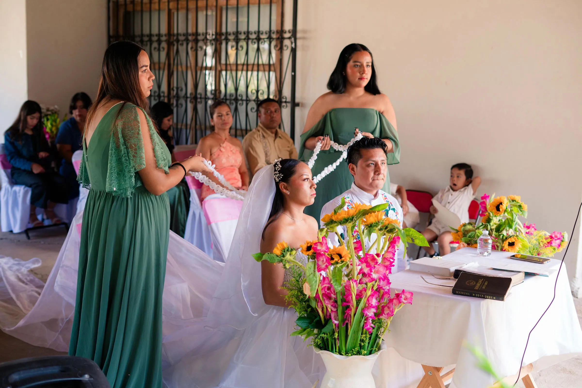 Two women in green dresses lower a white corded lazo over a seated bride and groom at a ceremony table decorated with sunflowers
