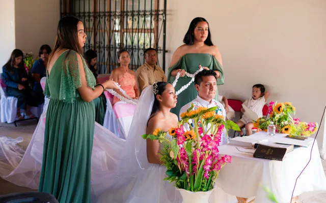 Two women in green dresses lower a white corded lazo over a seated bride and groom at a ceremony table decorated with sunflowers