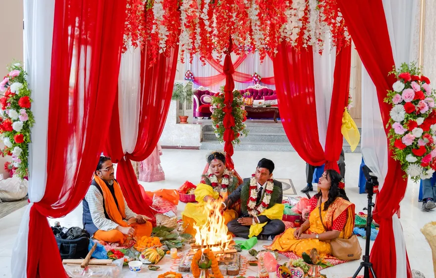 A mandap hung with red and white drapes and a canopy of red and white flowers, a small fire burning at its center while the couple and family sit on the floor around it