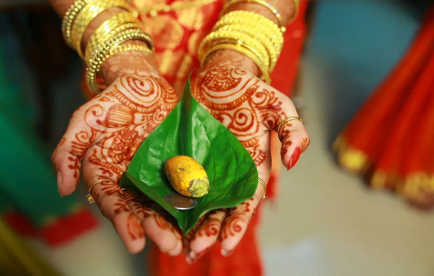 A bride's hennaed hands, stacked with gold bangles, cupped around a green betel leaf holding a piece of turmeric and a coin