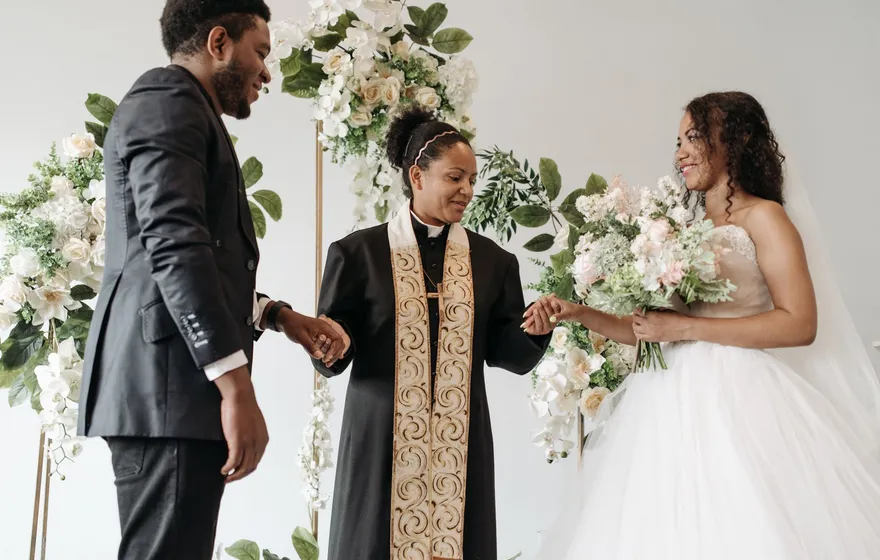 A Black officiant in a clerical robe and gold stole joins the hands of a bride in a white gown and a groom in a dark suit beneath a white floral arch