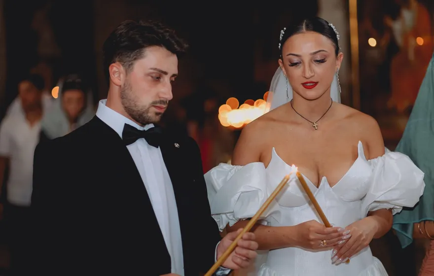 A bride and groom hold long lit tapers close together during a candlelit ceremony, guests blurred in the warm background