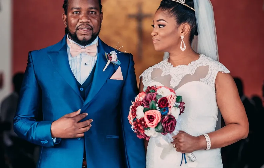 A groom in a bright blue suit and a bride in a white gown holding a red bouquet stand hand in hand in a church in Port of Spain