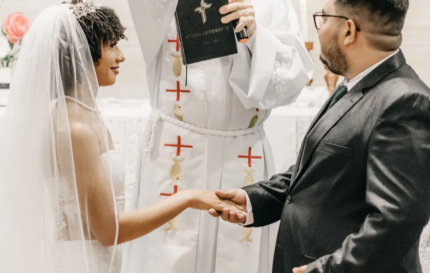 A gray-haired priest in a white chasuble raises his right hand in blessing over a bride in a veil and a groom in a dark suit who are holding hands