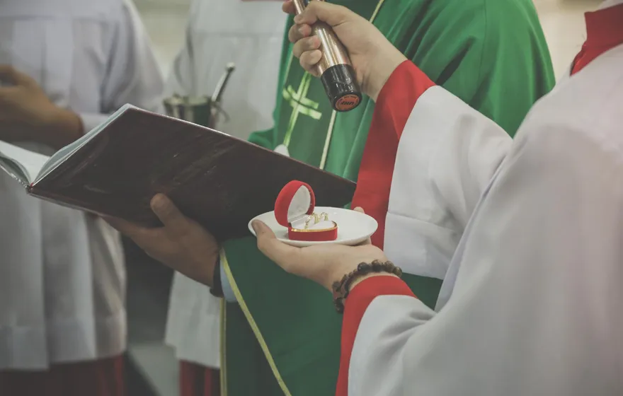 A priest in green vestments holds an open book while a server holds a small dish with a red ring box, an altar server in white and red standing beside them