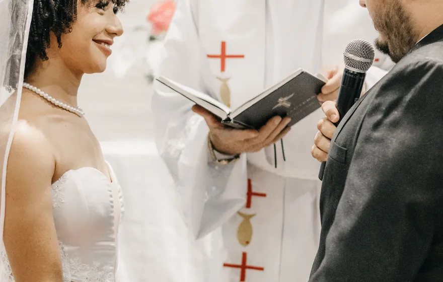 A priest in white vestments reads from an open book as a bride and groom hold hands in front of him, the groom speaking into a microphone
