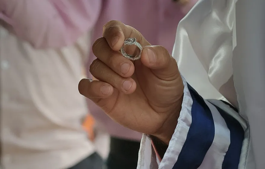 Close-up of a groom's hand holding up a braided silver ring, his sleeve wrapped in a white tallit with navy stripes