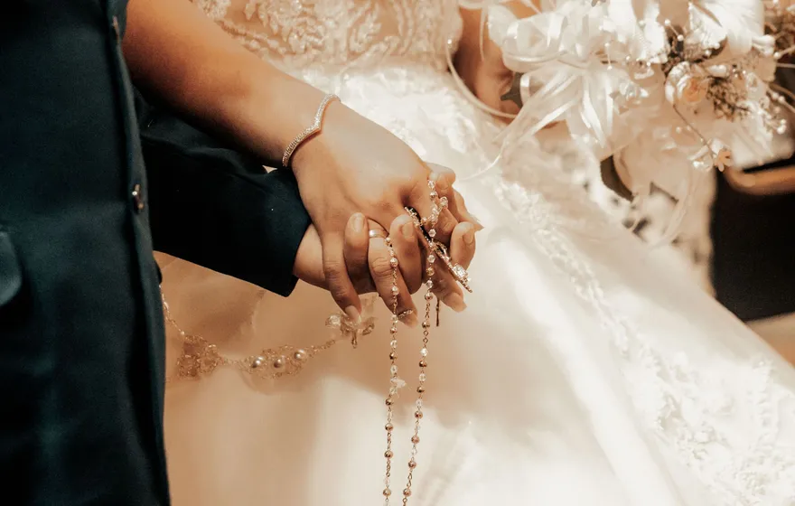 Close-up of a bride and groom holding hands over her white lace gown, a rosary wrapped through their fingers