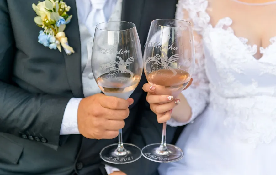 A bride and groom hold two etched wine glasses of pale pink wine side by side
