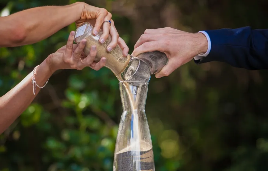 Two hands tip small jars of sand into a tall glass vase standing on a wooden barrel, greenery blurred behind