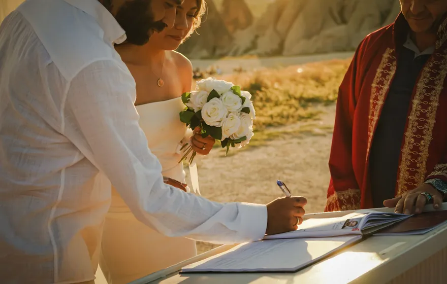 A groom signs a document on a table at an outdoor ceremony at sunset while the bride holds her bouquet and the officiant waits beside them