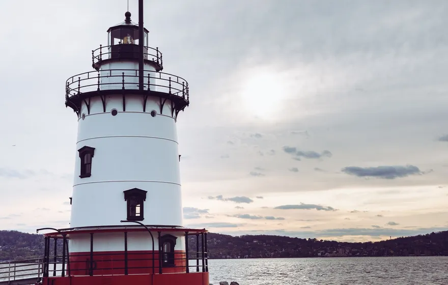 A white lighthouse with a red base stands in the Hudson River at dusk, a footbridge leading to it and wooded hills across the water