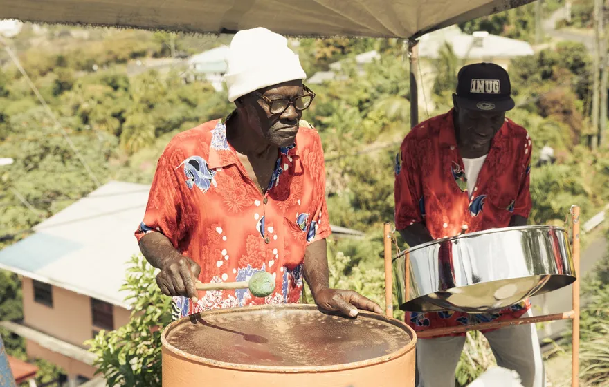 Two older men in matching red patterned shirts play steelpans under a canopy on a hillside in Trinidad and Tobago